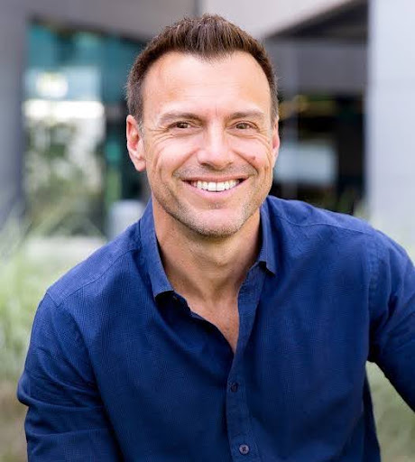 Smiling man in a blue button-down shirt outdoors, looking at the camera.