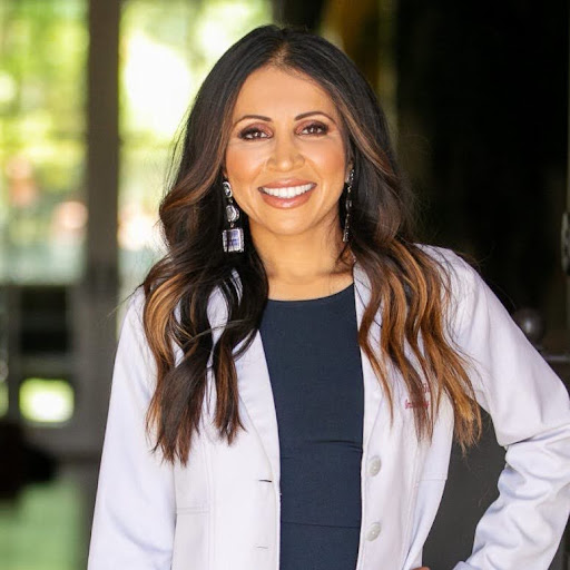 Smiling woman with long dark hair wearing a white lab coat and navy dress, standing outdoors near a glass building.
