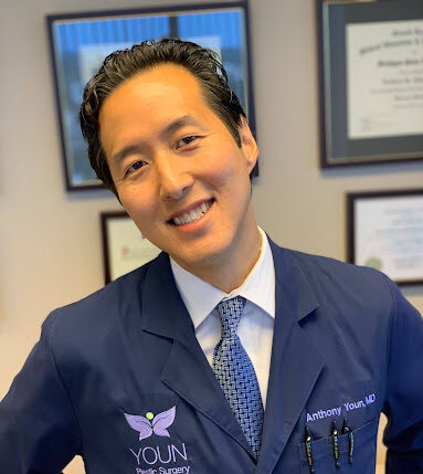 Dr. Anthony Your, MD, in a navy blue coat with YOUN Plastic Surgery logo, smiling in an office with framed certificates on the wall.