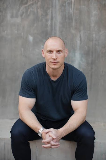 Casual portrait of a bald man in a blue t-shirt sitting on concrete steps, hands clasped, looking at the camera.