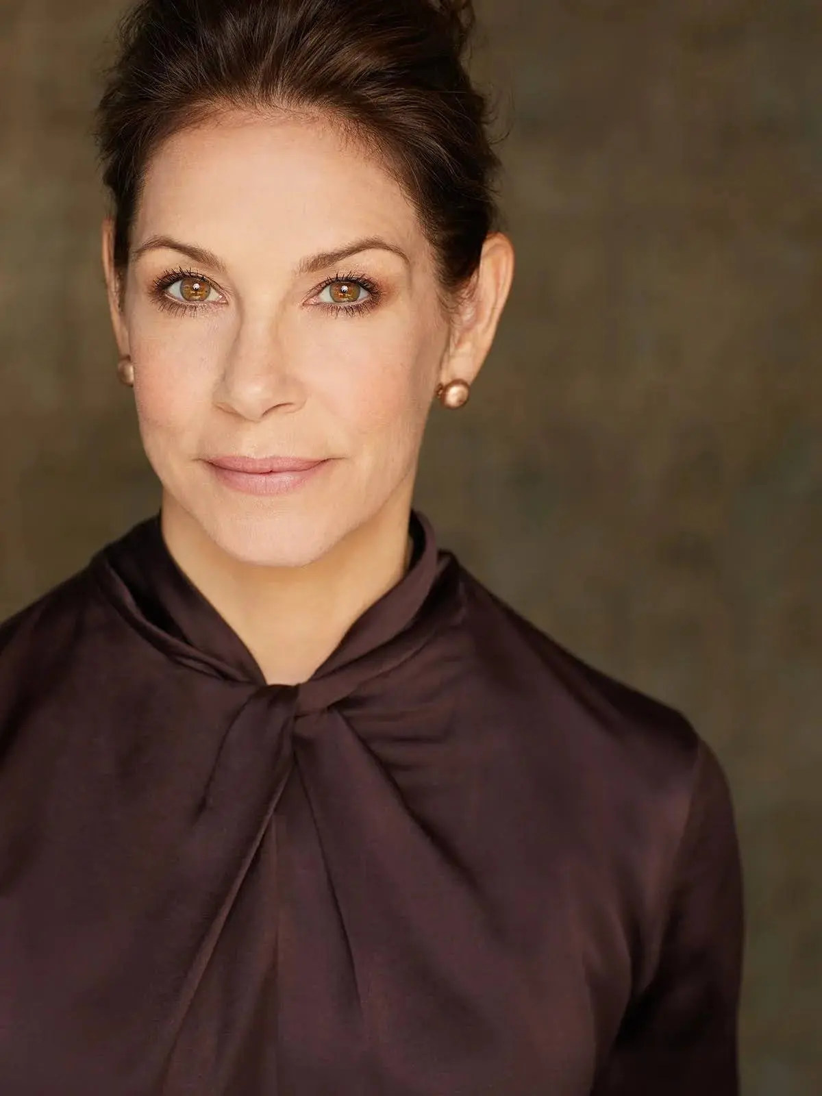 Head-and-shoulder portrait of a woman with brown hair, wearing a dark brown blouse with a bow, neutral background.
