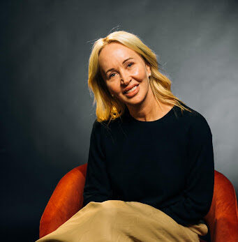 Smiling blonde woman in a black top sits on an orange chair against a dark studio backdrop.