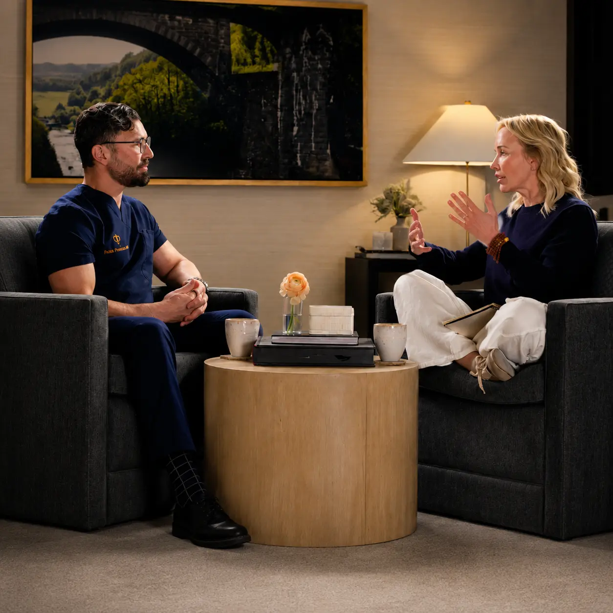 A man in scrubs and a woman in dark clothing sit on gray couches, talking in a cozy lounge with a round coffee table between them.